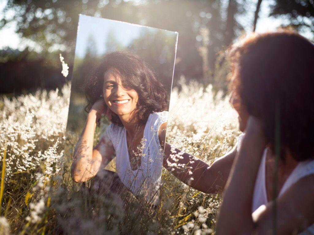 pexels photo 9917182 9917182 1 A woman smiles at her reflection in a mirror placed in a sunny field.